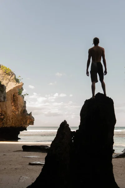 Die Silhouette eines jungen Mannes, der auf einem Felsen an einem Sandstrand steht. — Stockfoto