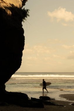 Sahilde sörf tahtasıyla yürüyen bir adam.
