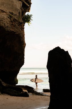 Sahilde sörf tahtasıyla yürüyen bir adam.