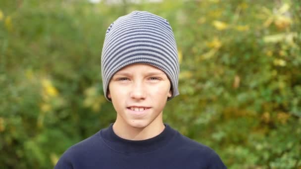 Улыбающийся мальчик в полосатой шляпе стоит в парке и смотрит в камеру. — стоковое видео