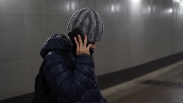 Eenzame jongen in hoed en jas praten over de telefoon in ondergrondse overdekte passage in de stad in de winter — Stockvideo