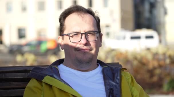 Πορτρέτο του όμορφου άντρα με γυαλιά που κάθεται σε παγκάκι στο πάρκο της πόλης, κοιτάζοντας με χαμόγελο στην κάμερα — Αρχείο Βίντεο