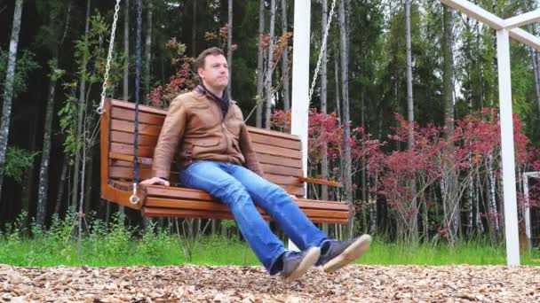 Millenium człowiek mówi na smartfonie siedząc na drewnianej ławce swinging w jesiennym parku na świeżym powietrzu — Wideo stockowe