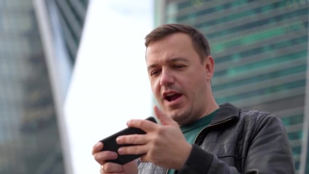 Emotionele geëxcideerde duizendjarige man spelen video game op de smartphone in de voorkant van moderne gebouwen — Stockvideo