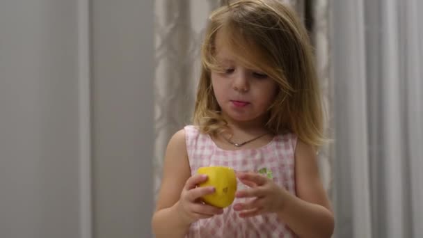 Portrait of cute baby girl in dress who eats a sour lemon while sitting indoors on high chair — Wideo stockowe