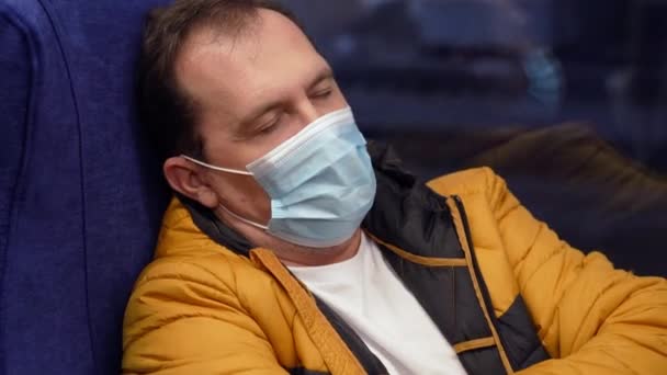 Κουρασμένος άνδρας με ιατρική μάσκα κάθεται κοντά στο παράθυρο του τρένου στην πόλη. Ο τύπος κοιμάται στις δημόσιες συγκοινωνίες. — Αρχείο Βίντεο