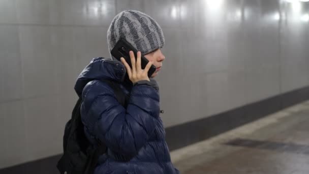 Lonely boy in hat and jacket talking on phone in underground covered passage in city in winter — Stock Video