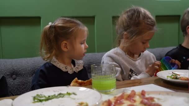 Kindermädchen spielen in buntem Anti-Stress-Spielzeug popit und kleines Kleinkind Mädchen isst Pizza im Café — Stockvideo