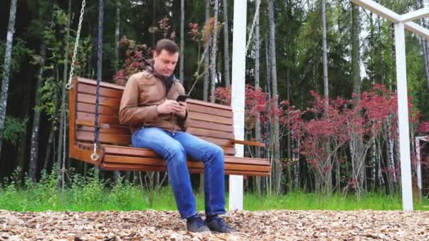 Guy texting on mobile phone in park. Young caucasian man relaxing on wooden swinging bench in autumn — ストック動画