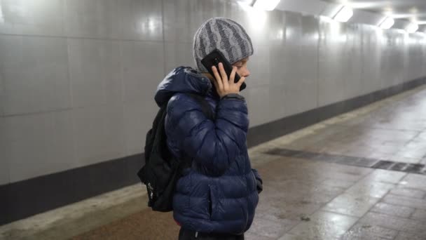Lonely boy in hat and jacket talking on the phone in underground covered passage in city in winter — Wideo stockowe