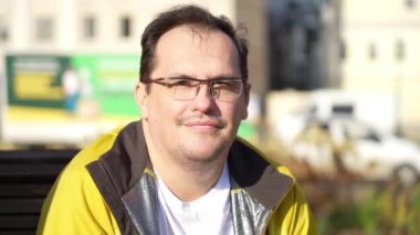 Good looking guy in glasses sitting on bench, relaxing in city park, looking with smile at camera