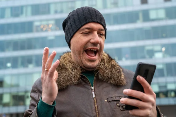 Excited guy in winter hat looks at phone and celebrates big win financial good news success achievement cell phone in front of modern buildings. Happy man checking news on smart phone walking in — Stock Photo, Image