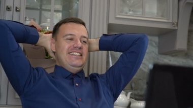 Millennial 'daki adam mutfakta dizüstü bilgisayarla rahatlıyor. Elleri başının arkasında oturuyor.