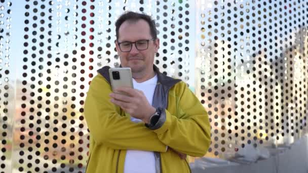 Hombre maduro en gafas apoyadas en la moderna pared metálica perforada gris en la ciudad, textos en smartphone — Vídeos de Stock