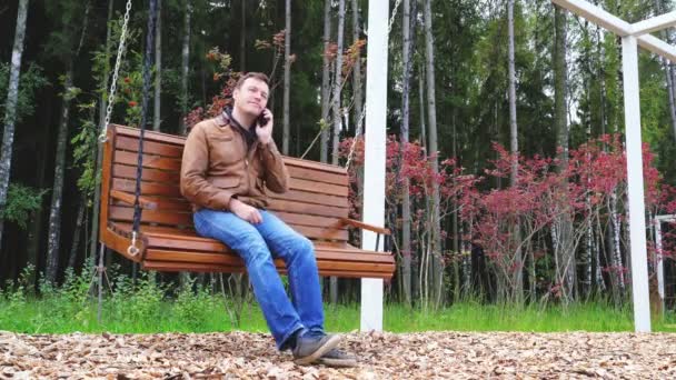 Joven hombre caucásico hablando en el teléfono inteligente mientras está sentado en el banco de madera oscilante en el parque de otoño — Vídeo de stock