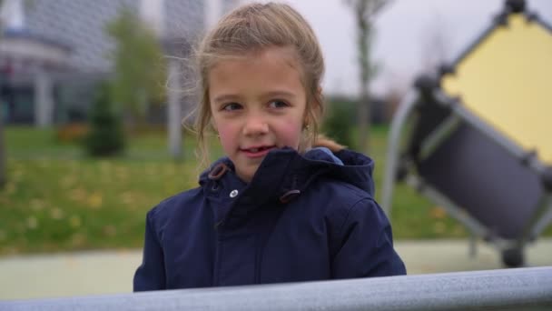Küçük kız kafasına örülmüş bir şapka takıyor ve kameraya bakıyor. Sonbahar parkında duruyor. — Stok video