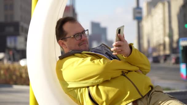 Adulto caucasiano homem relaxante, usando telefone celular em plástico branco balançando banco na primavera — Vídeo de Stock