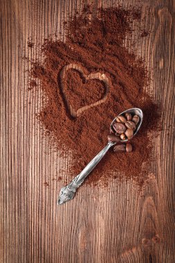 Metal kaşıkta kahve çekirdekleri ve tahta arka planda kalp şeklinde kahve tozu.