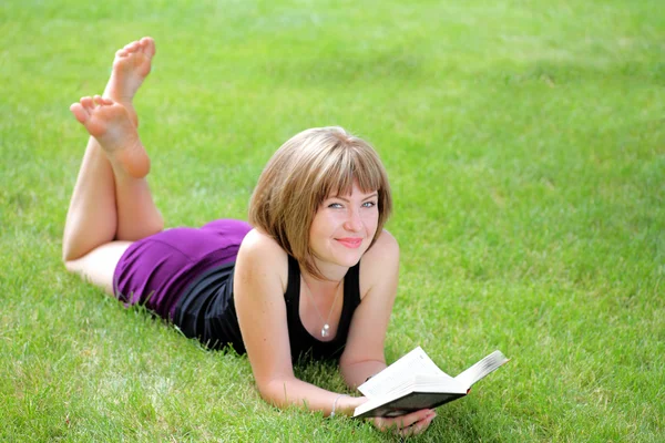 Gyönyörű fiatal mosolygó nő feküdt a füvön olvasó könyv — Stock Fotó