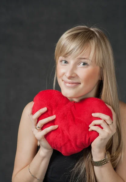 Beautiful young woman holding a heart shaped red pillow and smiling — Stock Photo, Image