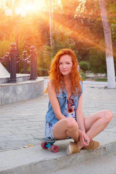 Red haired young women sitting on skateboard with her legs crossed backlit by sun — Stock Photo, Image