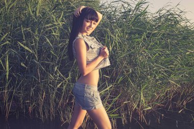 Sadece güneş ışığında kamışların önünde kot şortla poz veren esmer, zayıf kadın..