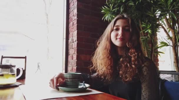 Гарна молода жінка коричневе волосся дивиться прямо на камеру під час пиття кави в кафе — стокове відео