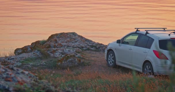 Unrecognisable car is standing on rocky sea cost with calm sea surface on background covered with orange sunset light. — Stock Video