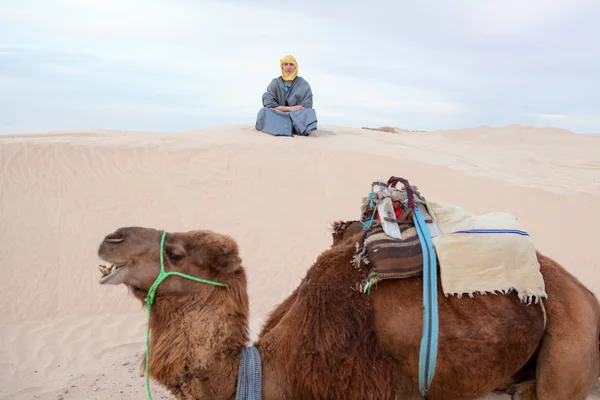 Caucasian woman sitting on sand dune in desert with camel on foreground — Stock Photo, Image