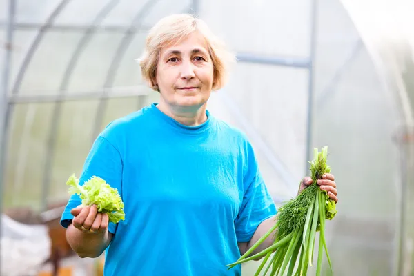 Feliz madura mujer caucásica con cebolla y lechuga delante del invernadero —  Fotos de Stock