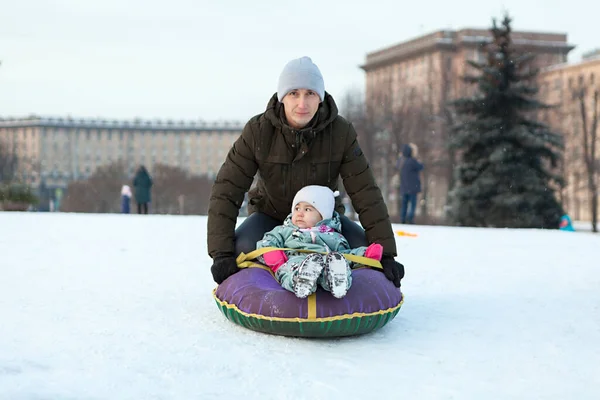 父は雪の斜面から彼女の子供をプッシュし 子供はインフレータブルトボガンに座って 冬の季節はロシアの都市にあります — ストック写真