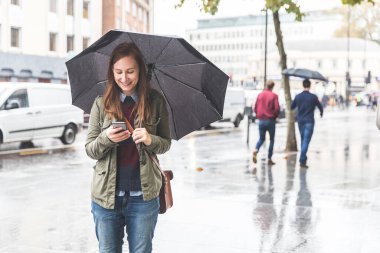 Londra 'da yağmurlu bir günde telefonunu kullanan mutlu bir kadın - gülümseyen, şemsiyesini tutan ve telefonla mesaj gönderen genç bir kadın - Londra' da hayat tarzı ve iş seyahati