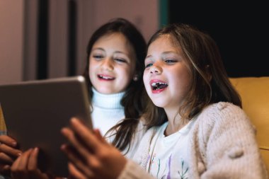 Evde dijital tabletle oynayan iki mutlu kızın sahnesinin sahnelendiği sahnedeki iki küçük kız kardeş kanepenin üzerinde oturmuş teknoloji aletini kullanarak gülüyor ve eğleniyorlardı.