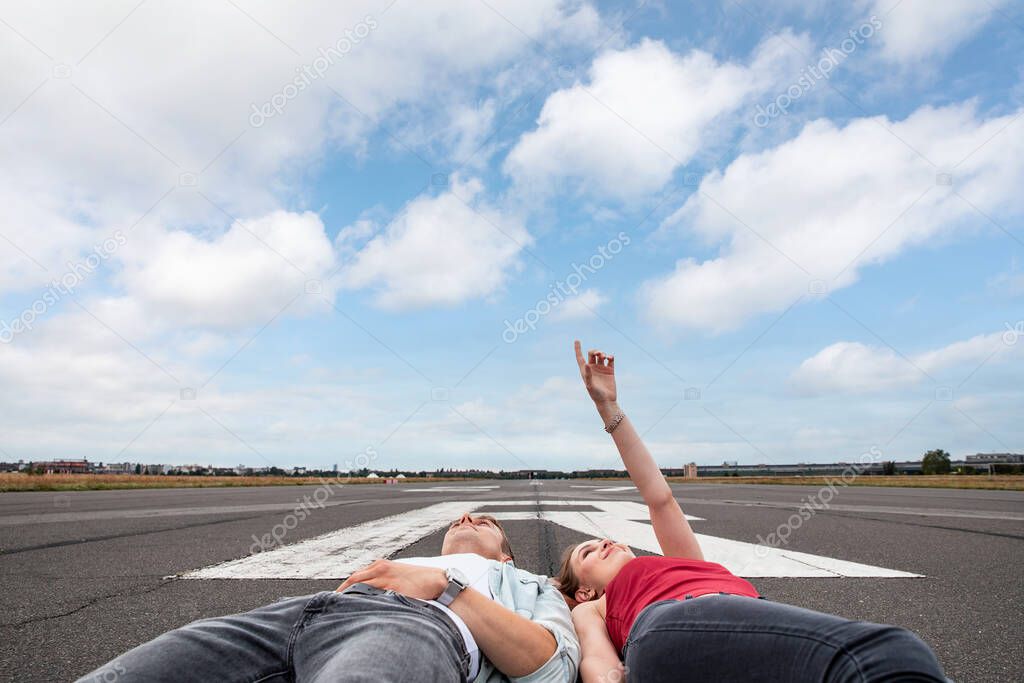 Happy couple lying down on airport runway and pointing at the sky dreaming of travel - Man and woman together planning beautiful life and future - transport and lifestyle concepts