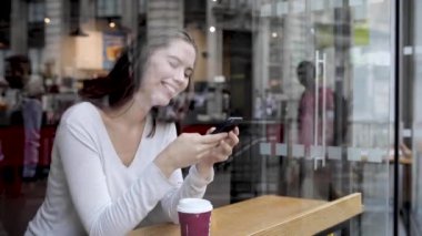 Bir kafede kahve içen ve akıllı telefon kullanan genç bir kadın Londra 'da bir kafenin penceresinde oturan güzel bir esmer, kameradan başka tarafa bakıyor - yaşam tarzı ve yiyecek içecek kavramları
