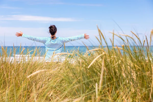 コーンウォールの海辺で海を見て腕を上げる女性 晴れた日に海を背景に砂丘に立つ女性の後ろの景色 旅行と幸福の概念 — ストック写真