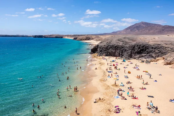 LANZAROTE, İspanya - 30 Haziran 2022: Deniz kenarında güneşli bir günde kristal berrak su ve beyaz kumların keyfini çıkaran insanlar