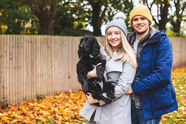 Happy couple and dog portrait at park with autumn leaves on the ground - Young couple holding a black dog and smiling at camera - Lifestyle and animals concepts