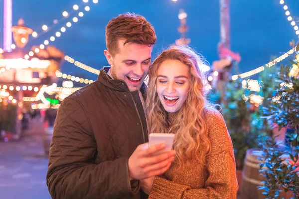 携帯電話を見て笑っている遊園地で幸せなカップル 楽しいフェアで夜を楽しんでいる若いカップル 愛と幸福の概念 — ストック写真