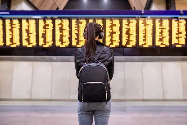 女性は駅で待っているとロンドンの到着出発ボードを見て 時刻表を見てヘッドフォンを持つ若い女性の後部ビュー コンピュータ旅行 ライフスタイル — ストック写真