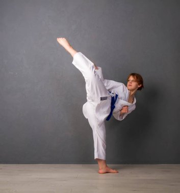 Beyaz kimonolu ve mavi kemerli genç bir kız, gri bir duvara karşı bir dizi egzersiz yapıyor.