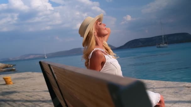 Ermioniの青い海の背景に木製のベンチに座って、太陽のわらの帽子の女性。ギリシャ — ストック動画