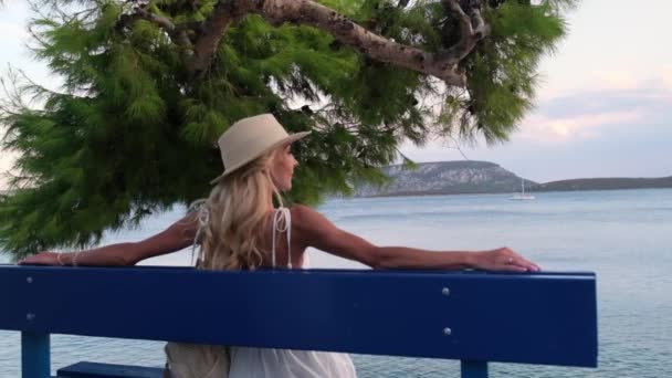 Mujer con sombrero de paja sentado en un banco con el mar y las montañas en el fondo, Ermioni, Grecia — Vídeo de stock