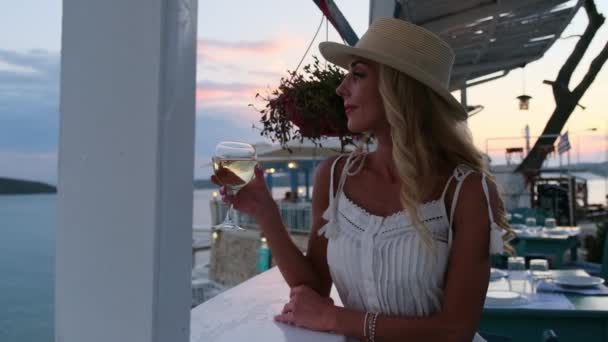Γυναίκα πίνοντας δροσερό λευκό κρασί στο υπαίθριο καφέ με τη θάλασσα και το ηλιοβασίλεμα στο παρασκήνιο — Αρχείο Βίντεο