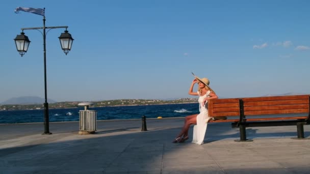 Vrouw in de zon strohoed zittend op een houten bank op blauwe zee achtergrond bij Spetses. Griekenland — Stockvideo