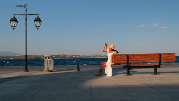 Vrouw in de zon strohoed zittend op een houten bank op blauwe zee achtergrond bij Spetses. Griekenland — Stockvideo