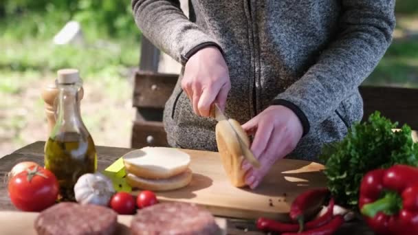 Женщина разрезала наполовину булочку с гамбургером ножом на деревянной доске на открытом воздухе — стоковое видео