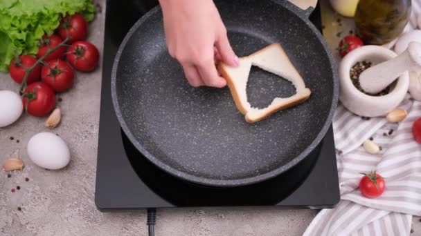 家庭のキッチンで熱々のフライパンに ハート型の穴が開いたトーストパンをかける女性 — ストック動画