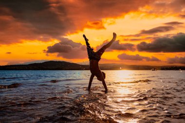 Çocuk gün batımında deniz kıyısında amuda kalkıyor.
