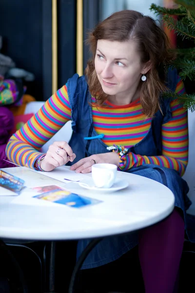 Beautiful girl in bright clothes writing postcards in cafe — Stock Photo, Image